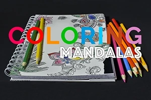 En ritbok med delvis färglagda mandala-mönster och färgpennor placerade ovanpå, med texten COLORING MANDALAS i flerfärgad och vit skrift
