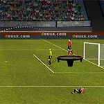 En fotbollsspelare i gul tröja sparkar en boll mot ett mål där en domare och en annan spelare står; bollen följer en kurvbana över planen
