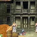 Två muskulösa män står framför ett gammalt övergivet hus med träfasad, omgivna av lådor, tunnor och en gammal bil