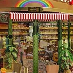 Exteriör av ett café med randig markis, regnbågsdekor och växter, inrett med korgstolar och bord